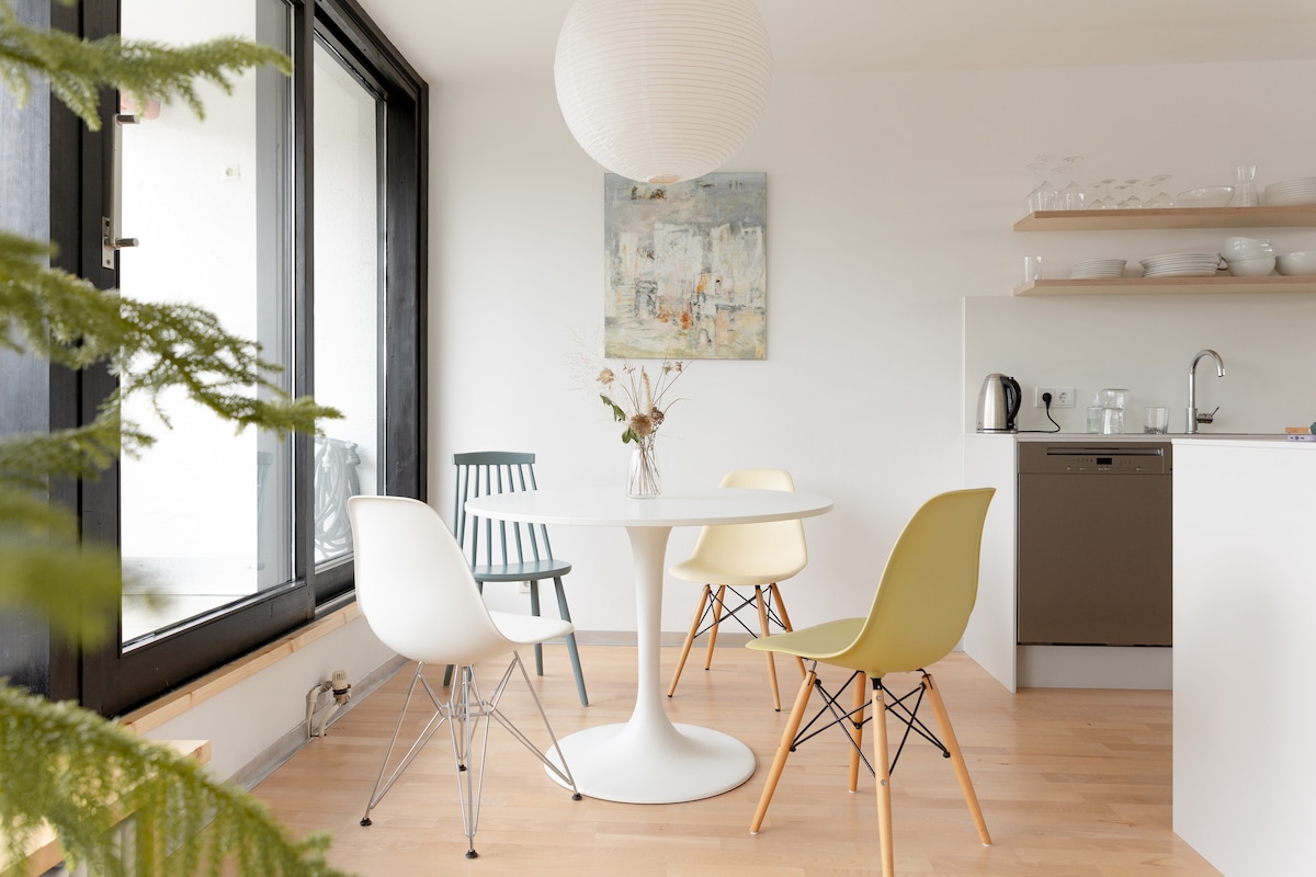 A dining area is presented with a round white table surrounded by four chairs in varying colors. Natural light enters through large glass doors, enhancing the bright and airy ambiance. A modern kitchen is visible in the background with minimalist shelving.
