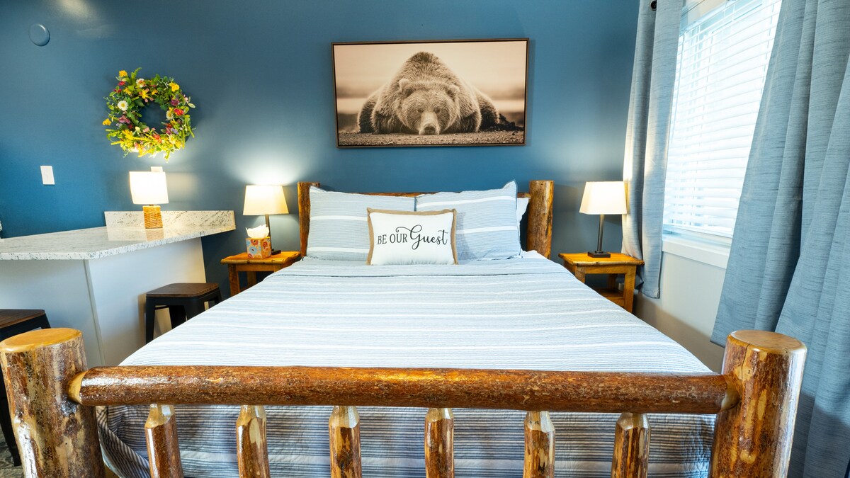 A spacious bed framed with rustic wooden posts is highlighted against a soft blue wall. A framed bear photograph hangs above the bed. Two bedside lamps illuminate the space, while a small kitchen area with marble countertop is partially visible. A wreath adorns the wall.