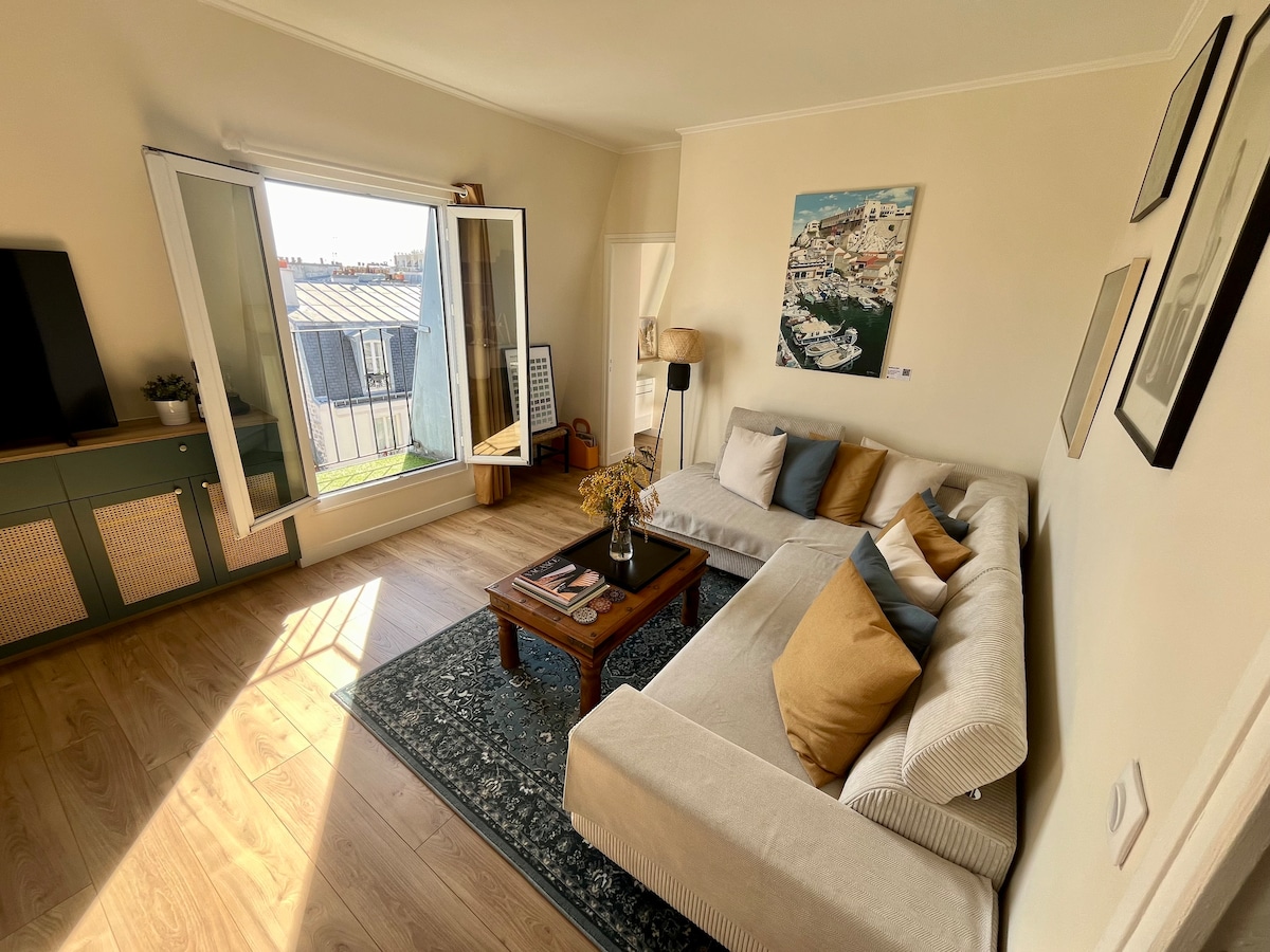 A spacious lounge area features a light-colored sofa adorned with cushions in various shades of blue and brown. A coffee table sits in front of the sofa on a patterned rug. Natural light floods the room through open windows, providing city views.