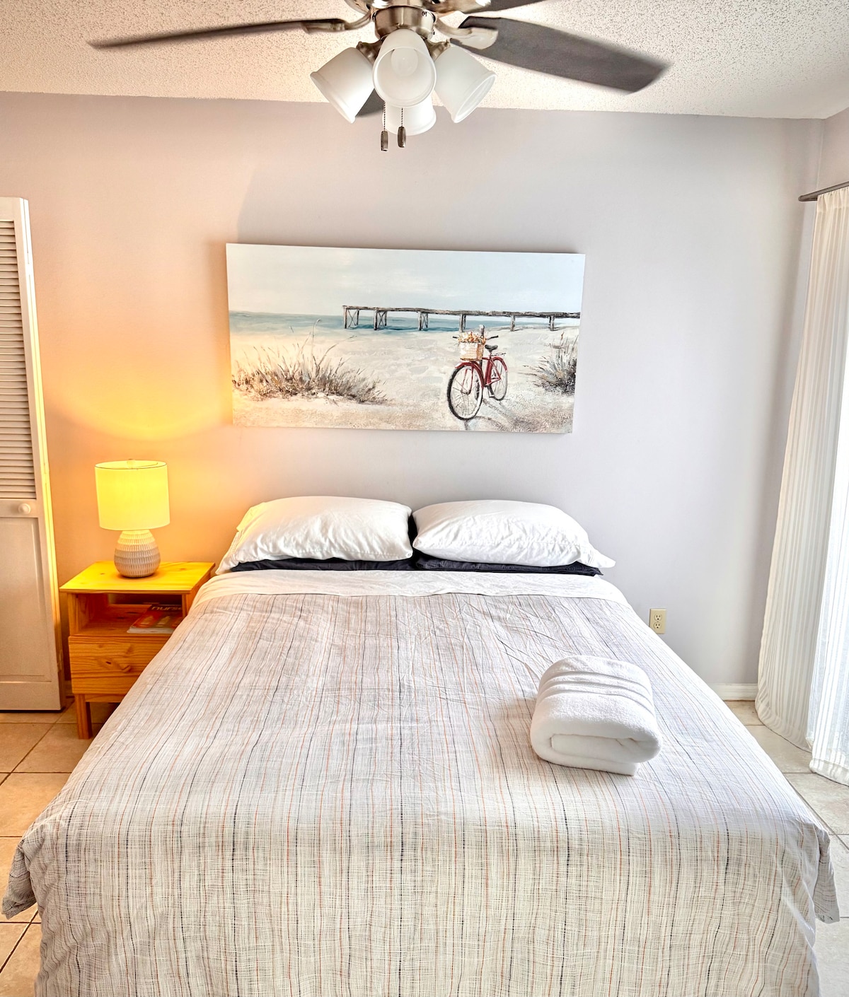 A cozy sleeping area features a spacious bed dressed in light linens. A large painting of a bicycle by the beach adds a serene touch to the room. A lamp on a wooden nightstand provides soft lighting, and natural light enters through sheer curtains.