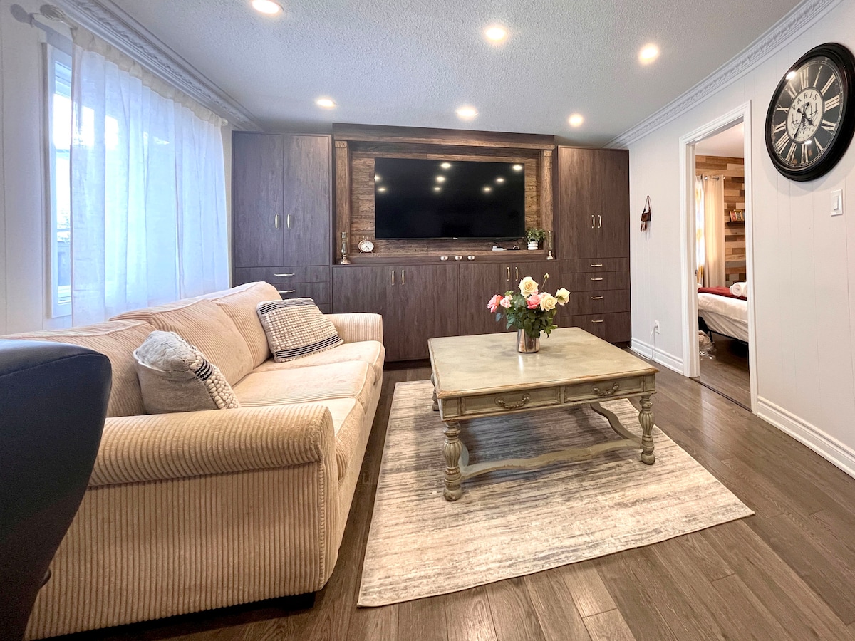 A comfortable living area is presented, featuring a beige sofa and a rustic coffee table at the center. A dark wooden entertainment unit accommodates a flat-screen TV, while soft lighting enhances the warm ambiance. Natural light enters through a window dressed with light curtains, illuminating the space.