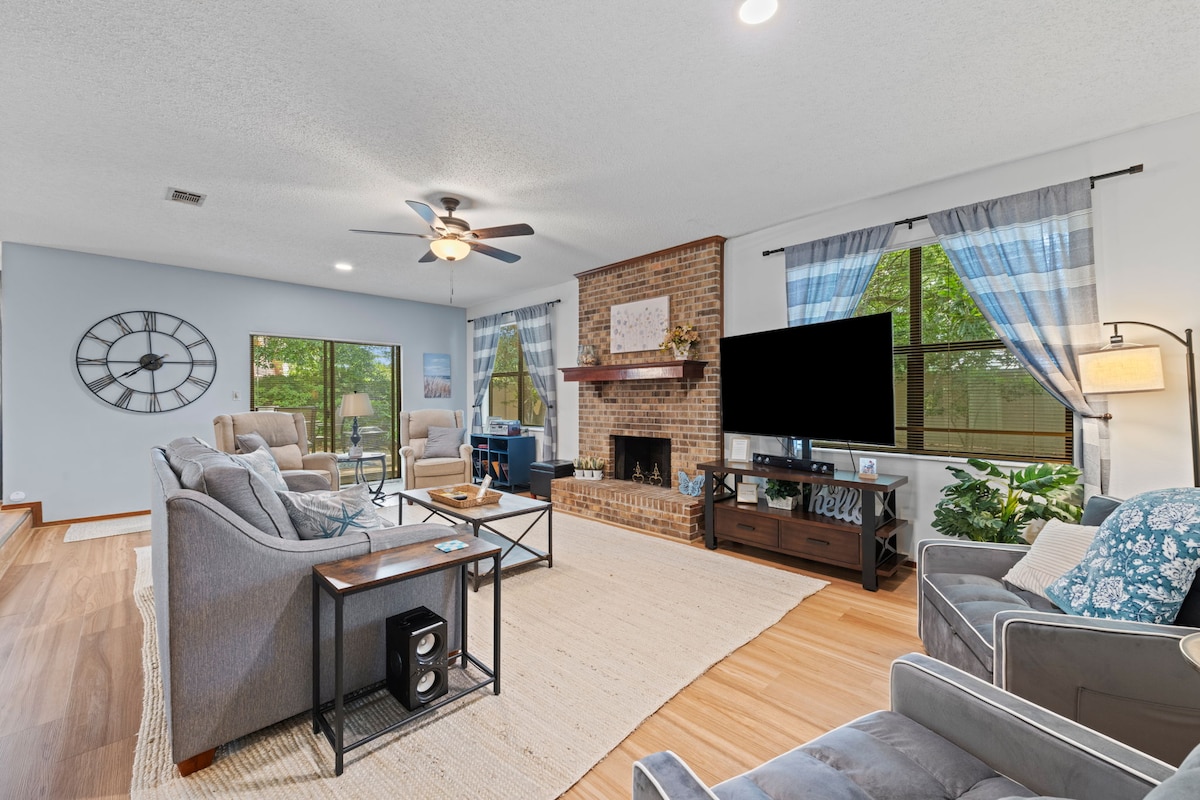 The spacious living room features a wood-burning fireplace and a large flat-screen TV. Comfortable seating includes a couch, two recliners, and two single sofa beds. Natural light streams through large windows with patterned curtains, illuminating the warm wood flooring.