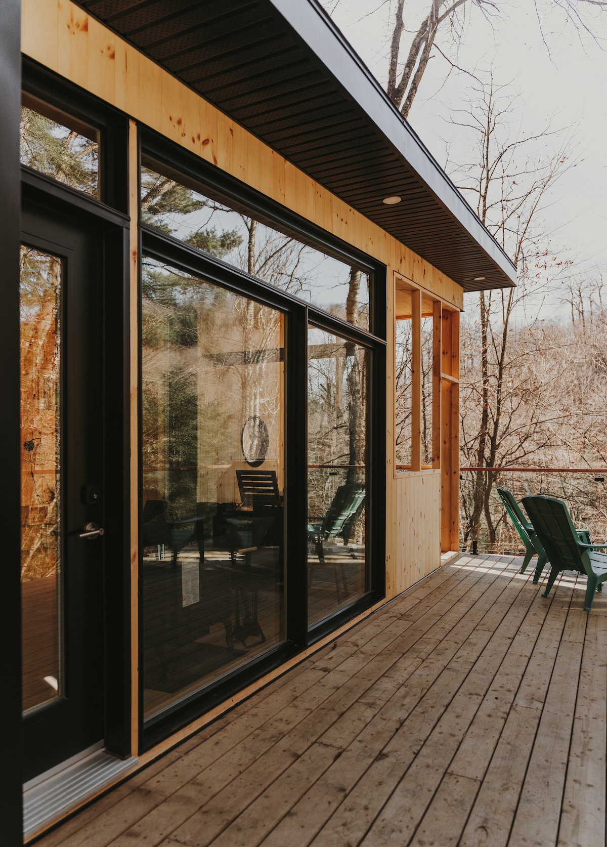 The image captures a modern exterior view of the chalet, highlighting large glass doors that connect the indoor and outdoor spaces. A wooden deck extends along the side, featuring several green lounge chairs. Surrounding trees offer a natural backdrop, enhancing the tranquil setting.
