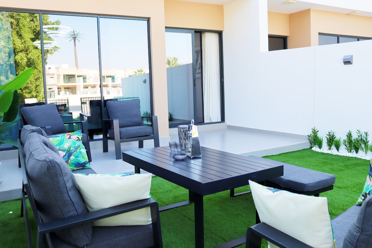 A spacious outdoor seating area features a low table surrounded by comfortable armchairs, all set on a lush green lawn. Large glass doors provide natural light and connect the space to the interior. Potted plants enhance the tranquil ambiance.