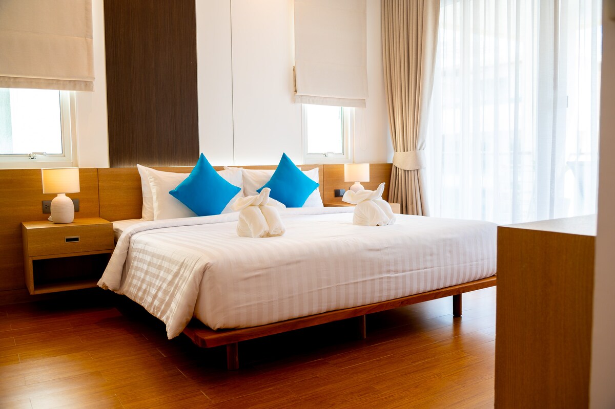 A spacious bedroom is illuminated by natural light from large windows. The king bed features crisp linens and decorative blue cushions. Minimalist wooden furniture and bedside lamps create a serene setting, complemented by the warm tones of the wooden floor.