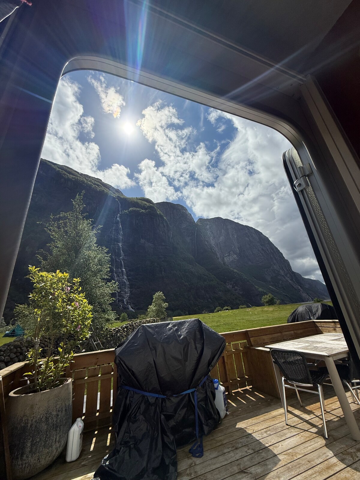 A scenic view is framed by the caravan's doorway, showcasing towering mountains and a waterfall cascading down rocky cliffs. Sunlight filters through the clouds, creating a bright atmosphere. An outdoor table and chairs are positioned on the spacious wooden terrace, surrounded by lush greenery.