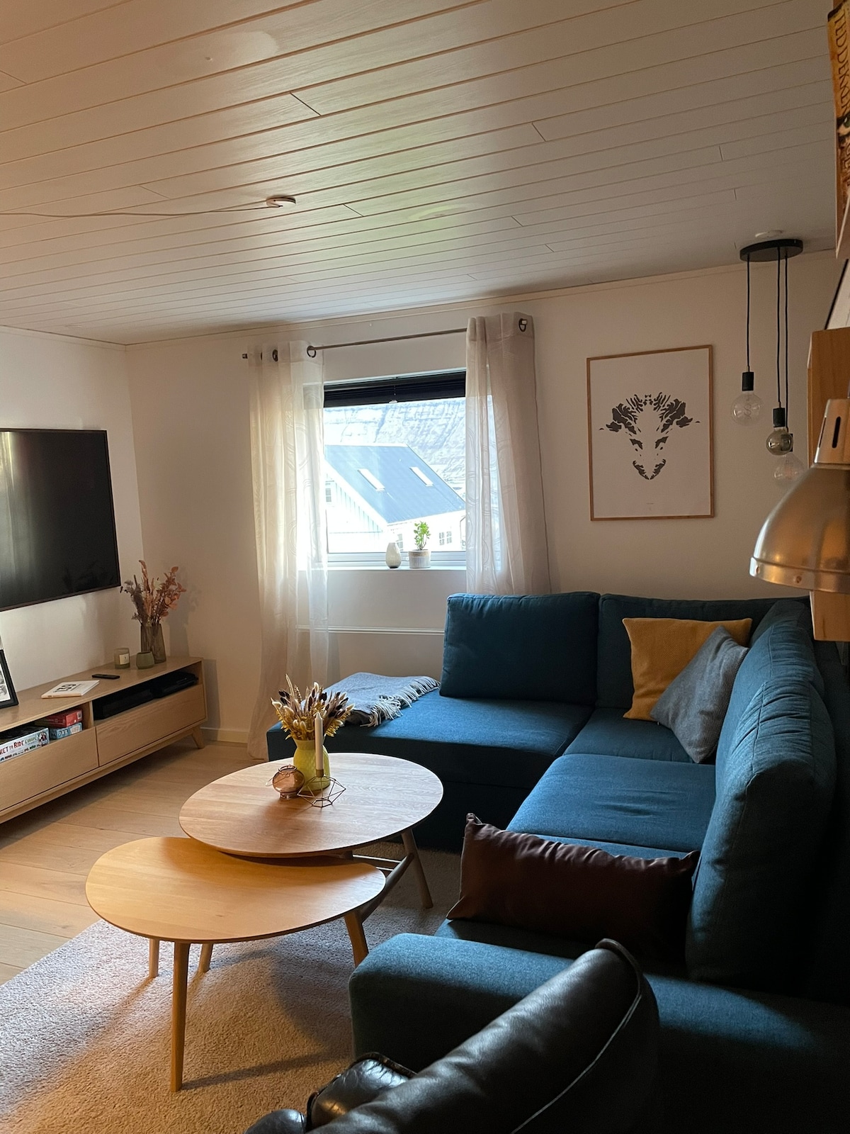 The living room features a cozy sectional sofa in a calming blue, accompanied by a round coffee table. A television is mounted on the wall, while light filters through sheer curtains, illuminating the space. An art piece is displayed on the wall, adding visual interest.