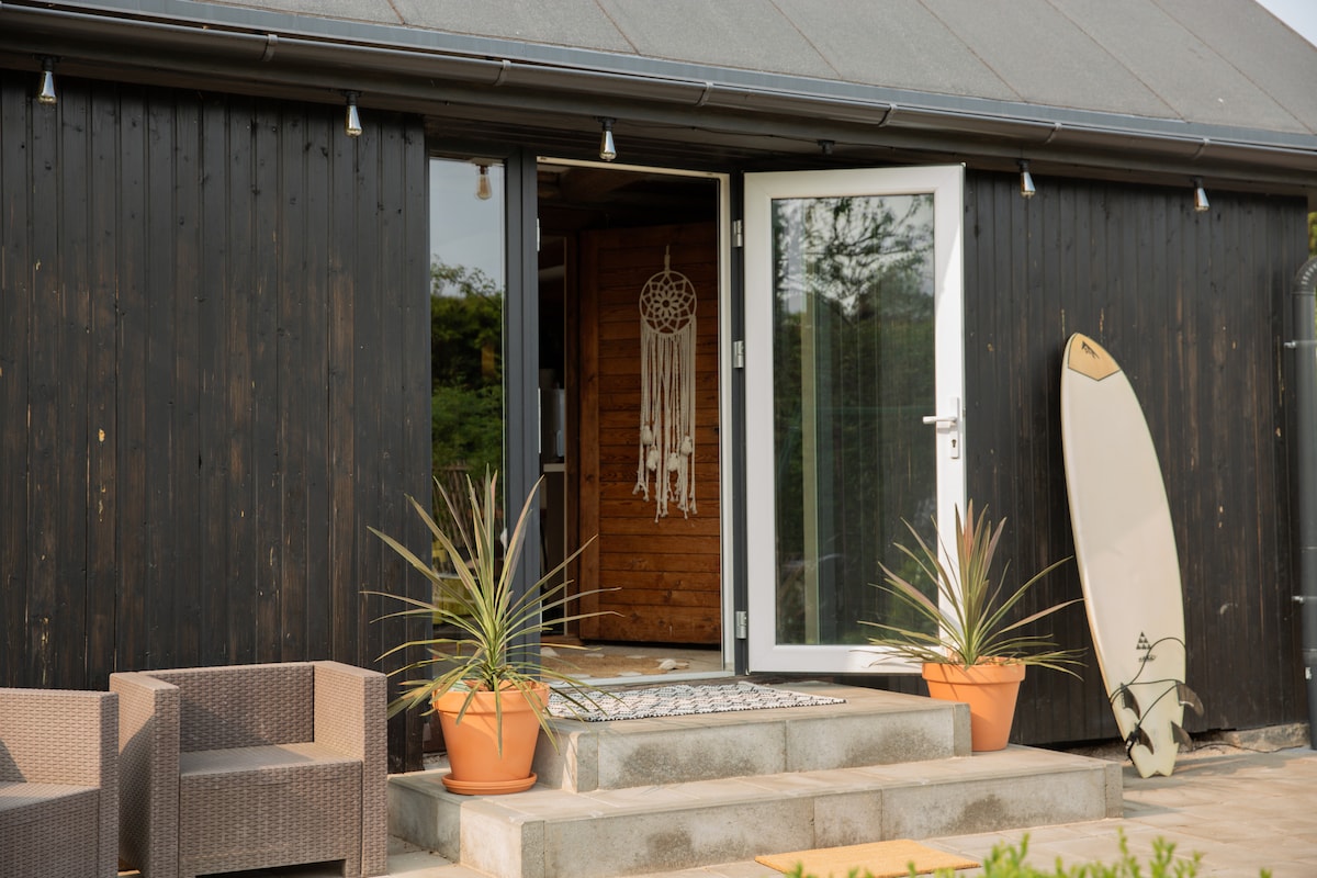 The entrance features a set of double doors, welcoming guests into the space. Potted plants are placed on either side of the steps, while a surfboard leans against the building. Two cozy seating arrangements are visible, enhancing the outdoor appeal.