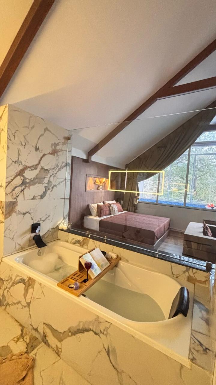 A spacious room is highlighted by a large window, allowing natural light to fill the space. A cozy seating area and a comfortable bed are visible, with a stylish bathtub featuring a wooden tray for relaxation. Elegant marble walls add a touch of sophistication.