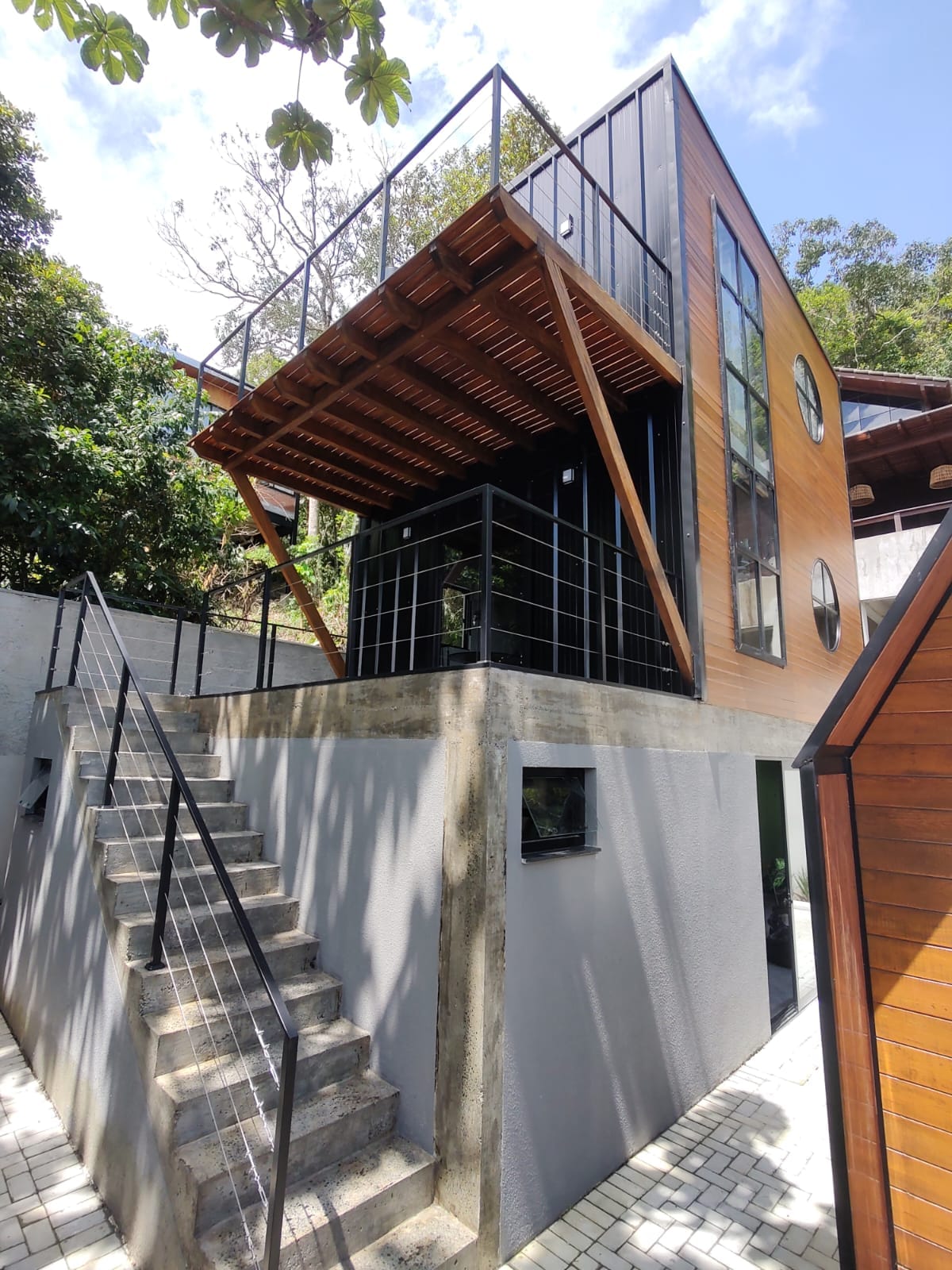 A modern triplex structure is showcased, featuring large glass windows and a wooden deck. Sturdy stairs lead to the upper level, surrounded by lush greenery that enhances the home's natural setting.