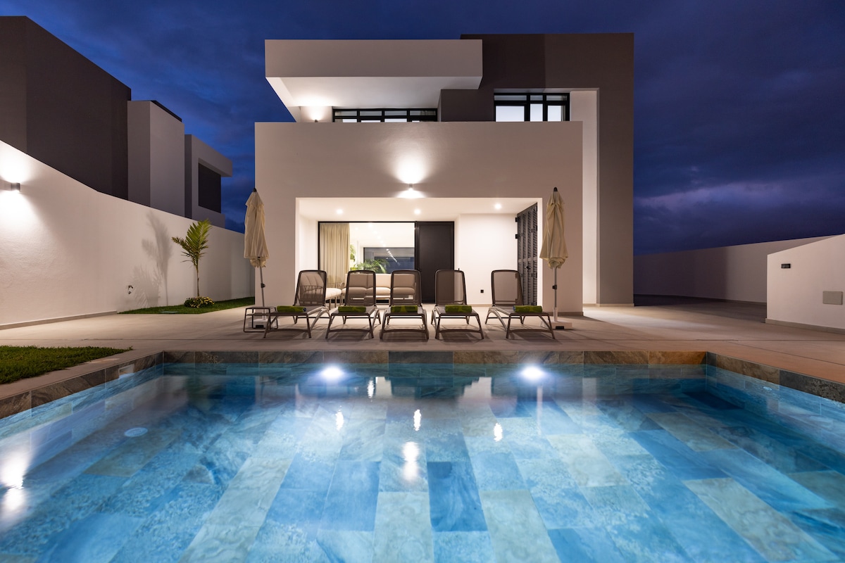 The modern house is depicted in twilight, featuring a private pool with clear blue water illuminated by built-in lights. Comfortable lounge chairs are arranged nearby on a spacious patio, and the structure showcases large windows that blend with the evening sky.