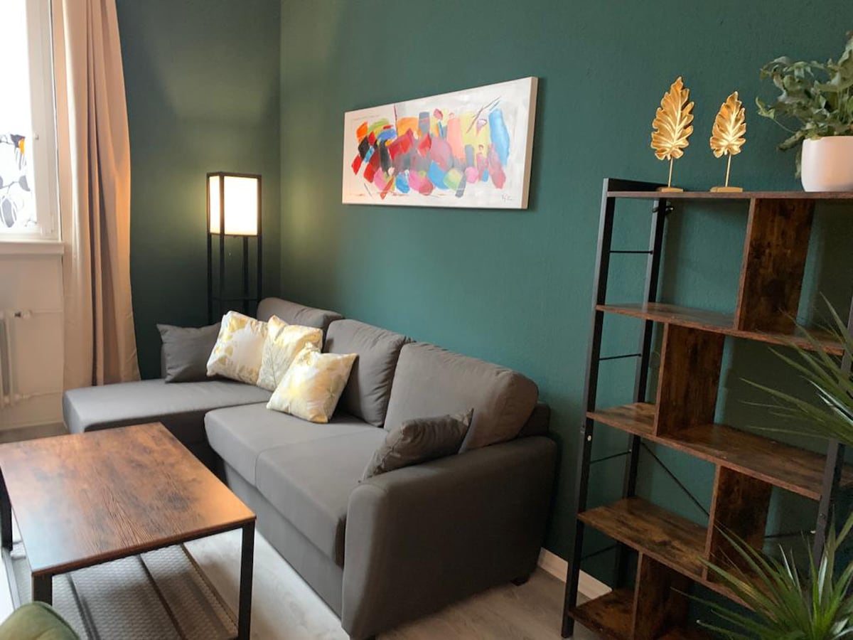 A modern living area features a gray sofa adorned with decorative pillows, inviting relaxation. A wooden coffee table rests in front, complemented by a minimalist shelving unit displaying plant accents and decorative items. Colorful artwork graces the wall, enhancing the room's visual interest.