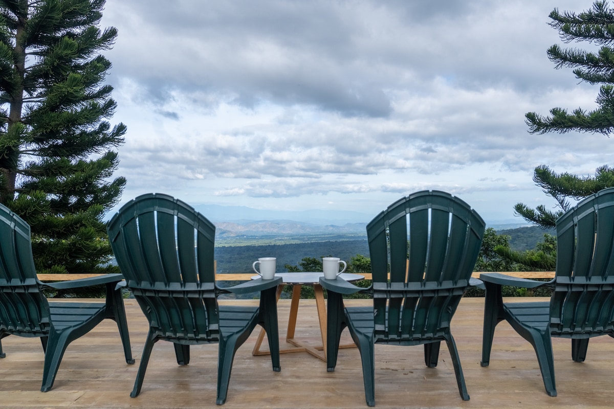 A spacious outdoor deck features four dark green Adirondack chairs arranged around a small wooden table. Two cups sit on the table, overlooking a breathtaking mountain view under a partly cloudy sky.