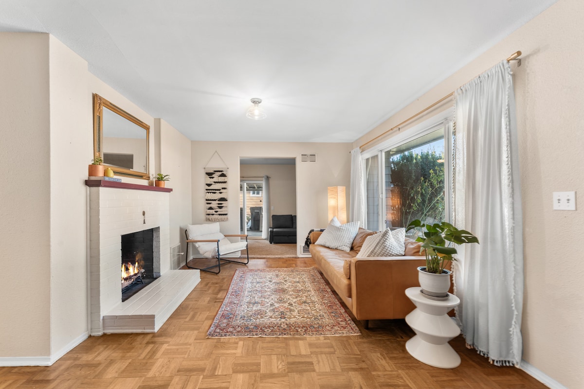 A cozy living room features a warm fireplace with a white brick surround and a large window allowing natural light. A brown sofa is paired with a patterned rug and stylish side tables. Adjacent seating is visible, creating a welcoming environment.