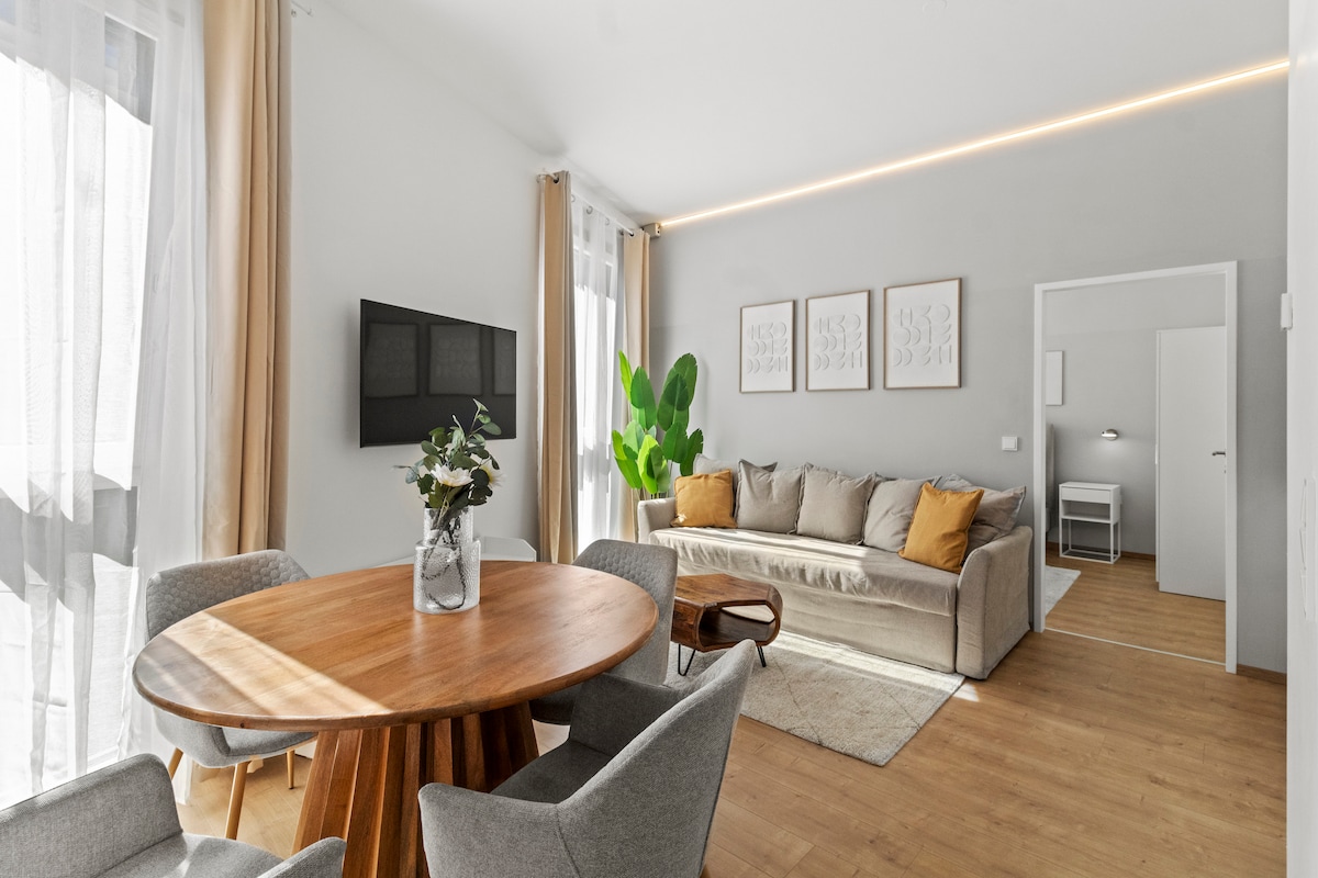 A light-filled living area features a round wooden dining table surrounded by four grey chairs. A spacious sofa with accent cushions is positioned against a wall, complemented by a decorative plant nearby. Large windows allow natural light to illuminate the room.