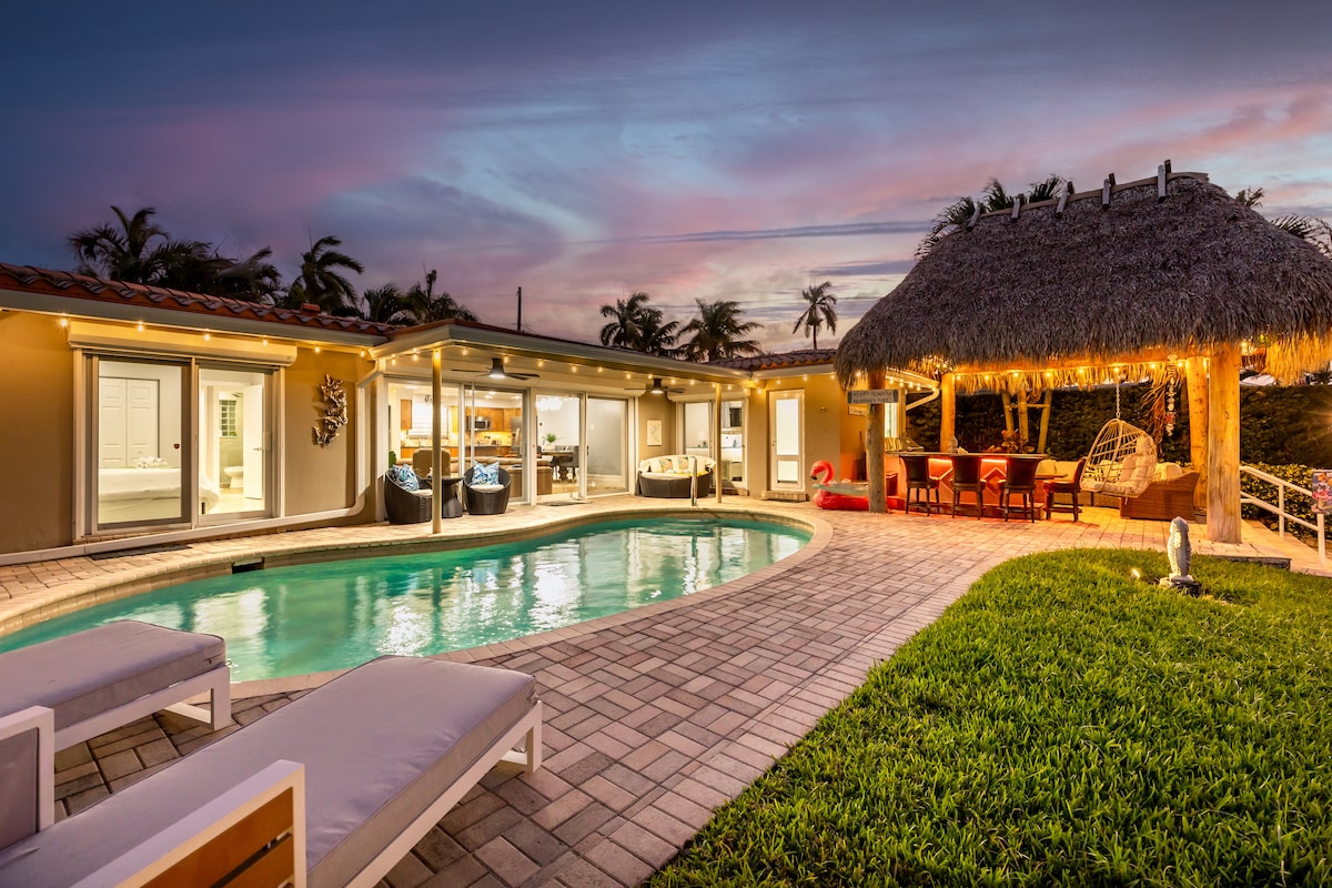 The outdoor area showcases a private pool surrounded by lounge chairs on a stone patio. A covered Tiki lounge adorned with lights provides seating, enhancing the inviting space. Lush greenery frames the area as evening light casts a warm glow across the scene.