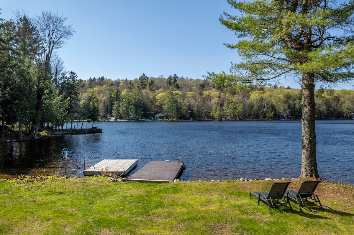 A scenic view of a tranquil lake is captured, featuring a wooden dock extending into the water. The green lawn is complemented by two black lounge chairs positioned nearby. Lush trees line the opposite bank, showcasing the natural beauty of the surrounding landscape.