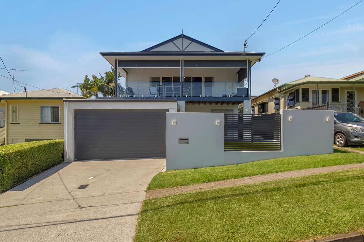 A modern, two-story house is presented with a spacious front yard and a concrete driveway leading to a lockable garage. The upper level features a covered balcony with glass railings, providing an area for outdoor relaxation, while the front facade showcases a contemporary design.