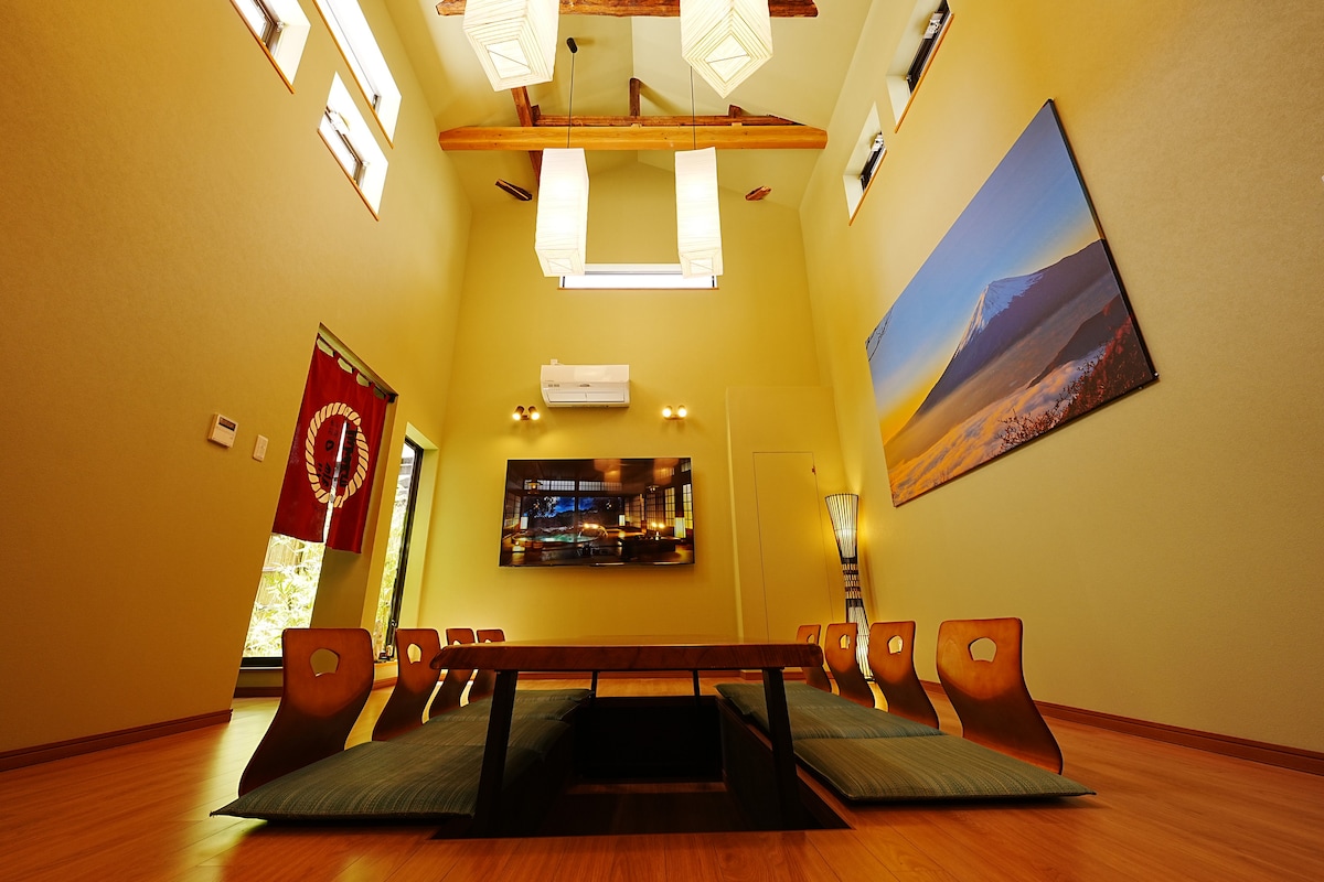 A spacious interior is highlighted by high ceilings and wooden beams. Tatami mats cover the floor, surrounded by low wooden seating. A large television is mounted on the wall, while soft lighting fixtures provide illumination, creating a serene environment.