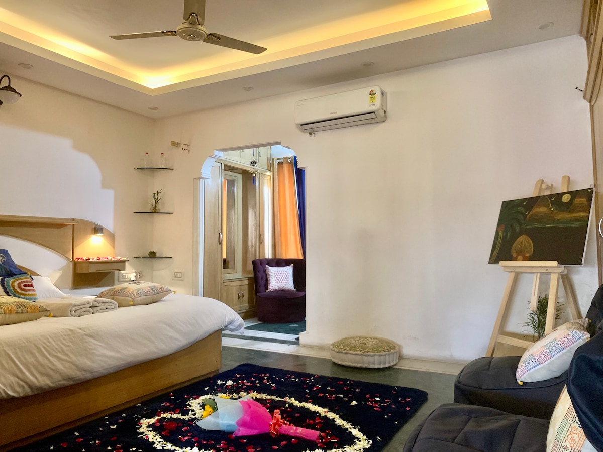 A serene bedroom is highlighted by soft lighting and a cozy bed adorned with decorative pillows. A plush armchair and a small side table are positioned nearby, while a colorful area rug anchors the space. Natural light flows through the doorway leading to a vibrant living area.
