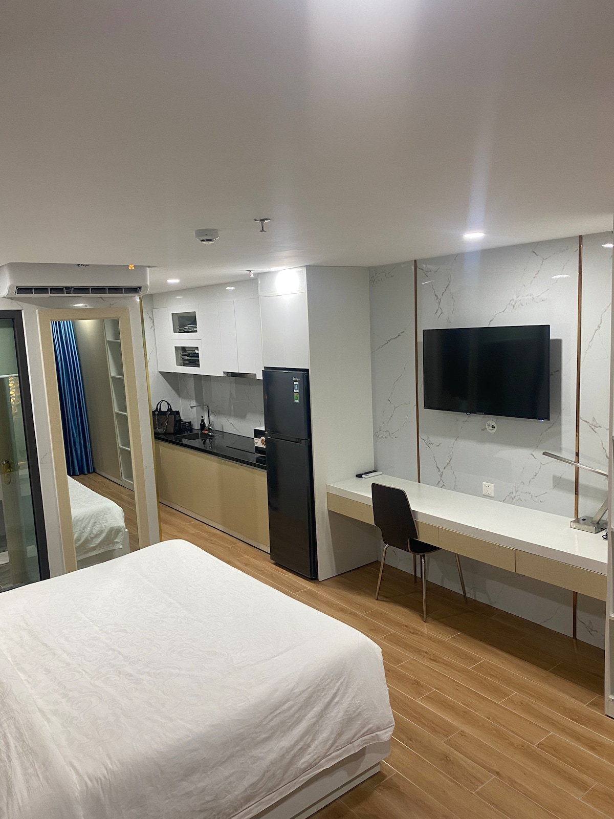 A modern living space is depicted, featuring a cozy sleeping area with a white bed covering. A compact kitchen area is visible, equipped with a refrigerator and a sleek black counter. A wall-mounted television is positioned above a light-colored desk, complemented by a dark chair.