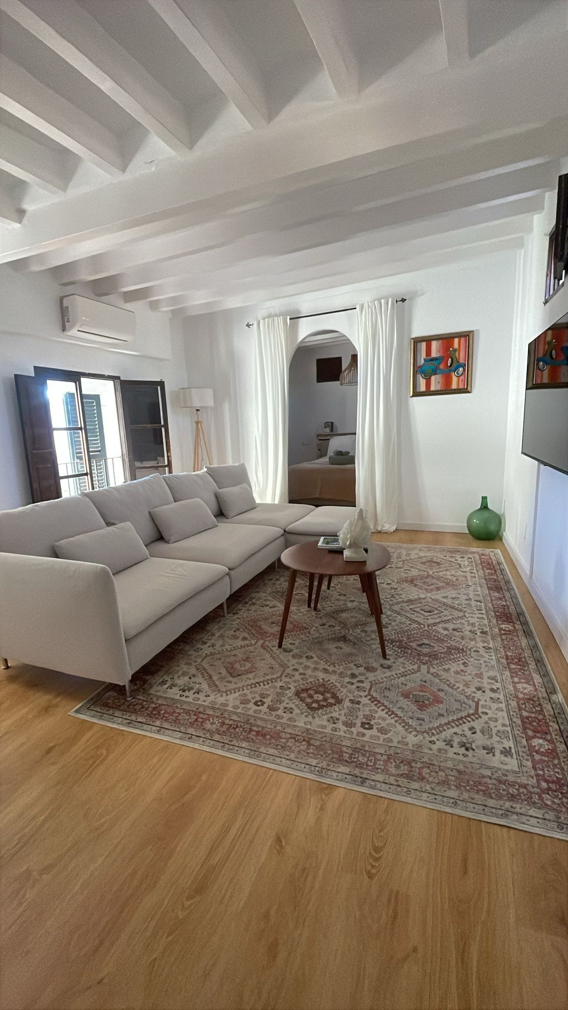 A bright living room is depicted, featuring a spacious light-colored sofa positioned against a wall. A small round table sits in front of the sofa. Natural light filters through the open window, illuminating the decor, which includes wall art and a green decorative bottle.
