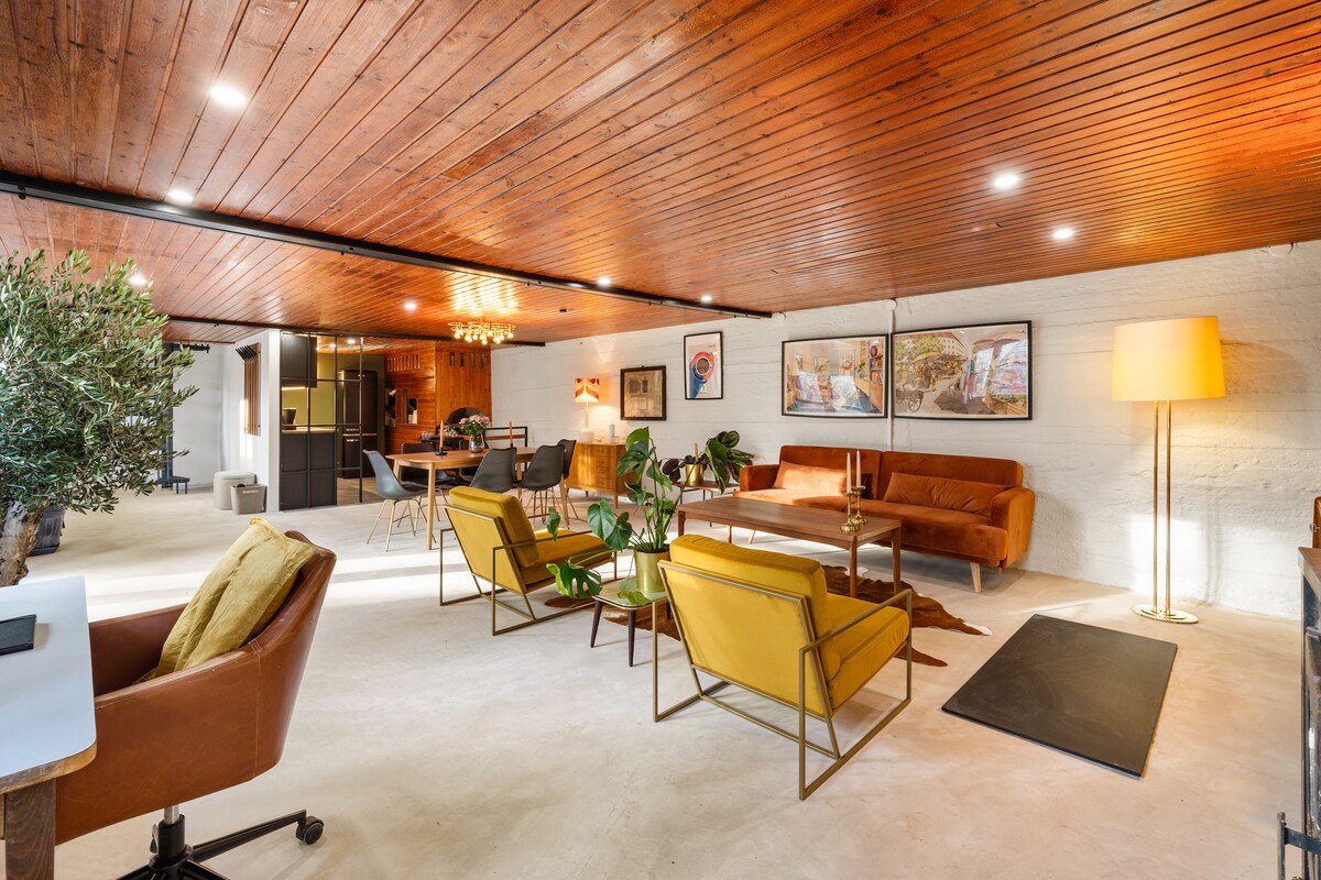 A spacious living area features warm wooden beams overhead and a light-colored floor. A vibrant orange sofa is paired with yellow chairs, complemented by decorative plants. A dining area is seen in the background, with a well-lit kitchen on one side.