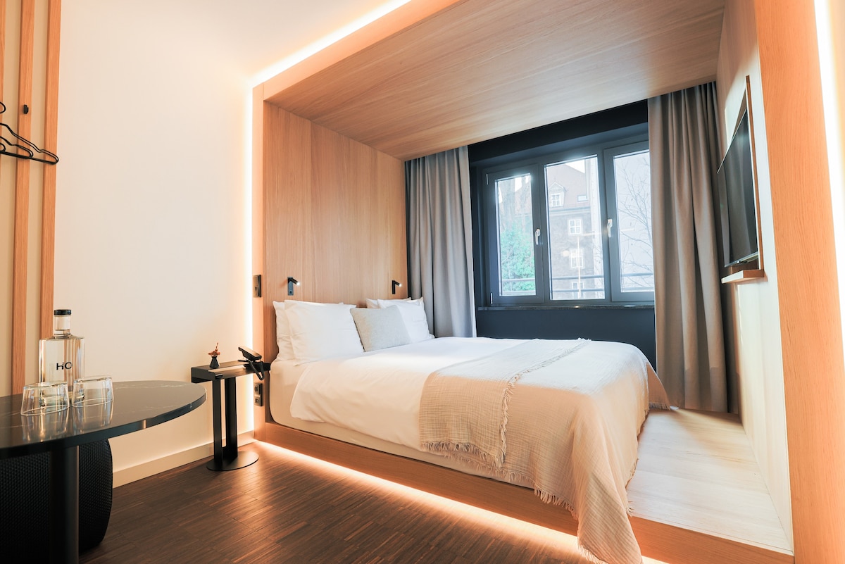 A minimalist bedroom features a spacious bed with crisp white linens and subtle textured accents. Natural light enters through large windows dressed with smooth, light grey curtains. A sleek table and modern lamp complete the warm wooden aesthetic of the interior.