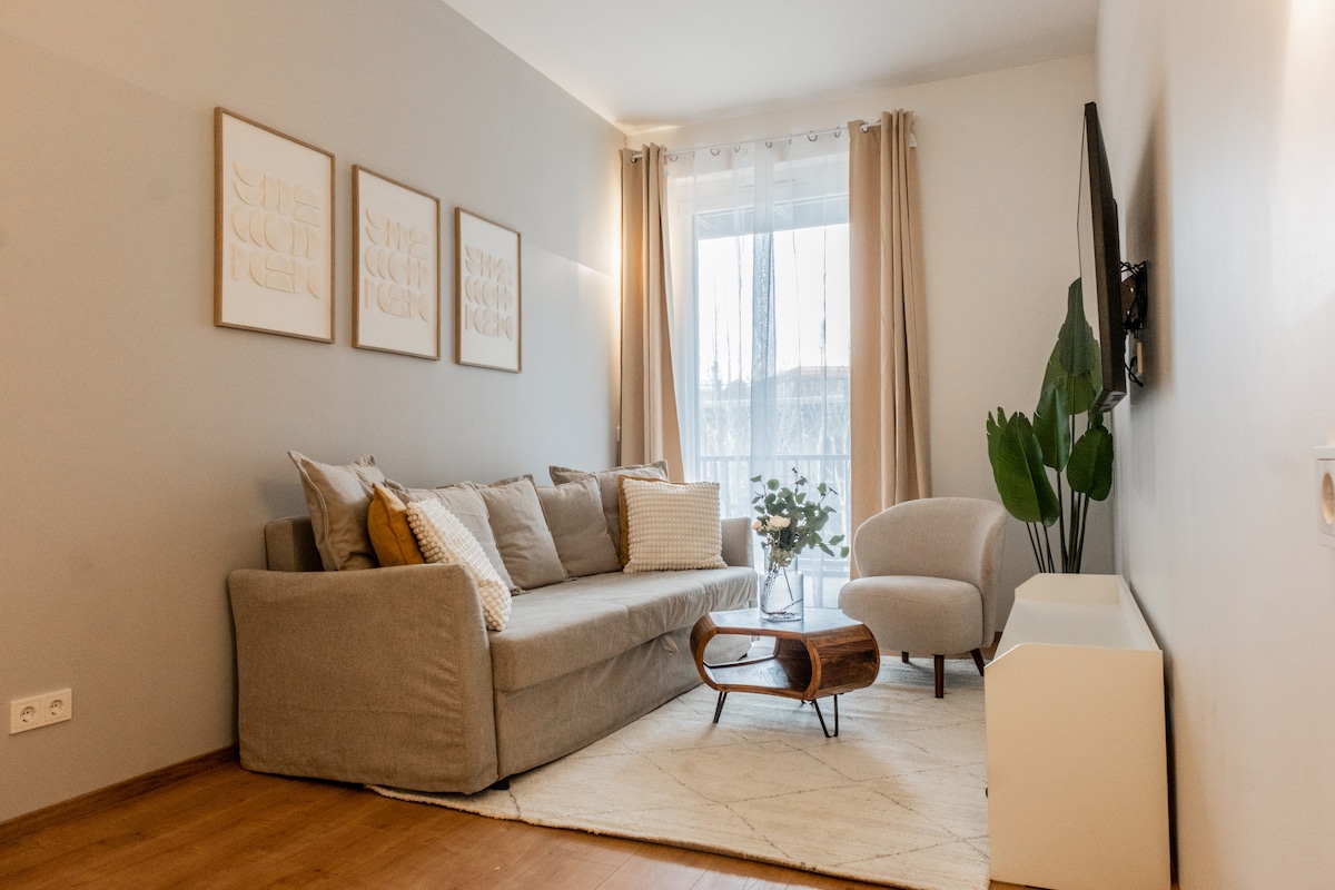 A comfortable living room features a cozy sofa adorned with decorative pillows, accompanied by a minimalist coffee table. Large windows allow natural light to fill the space, which is complemented by neutral-toned walls and light-colored curtains. Potted greenery adds a touch of nature.
