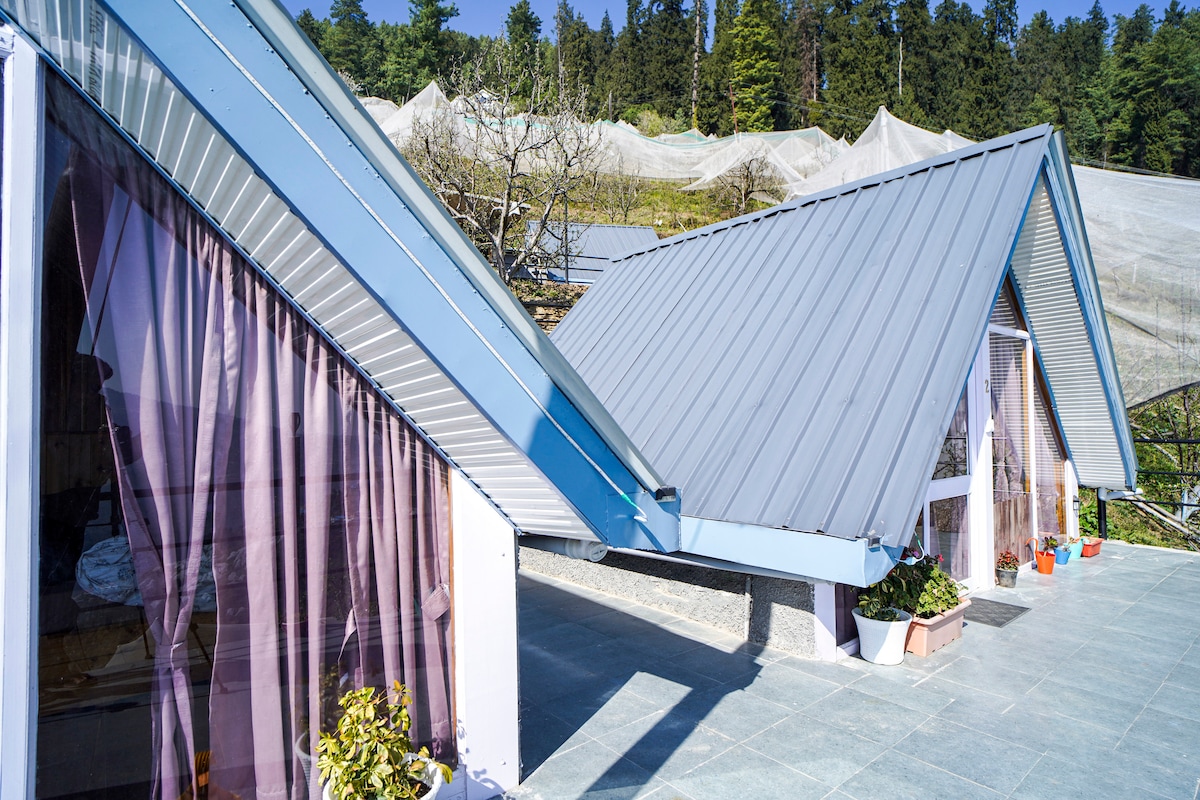 The image shows the exterior of the A-frame cottage with a sloped metal roof and large glass doors. Sheer curtains in soft hues can be seen fluttering in the breeze, while pots with plants adorn the patio. Lush greenery is visible in the background.