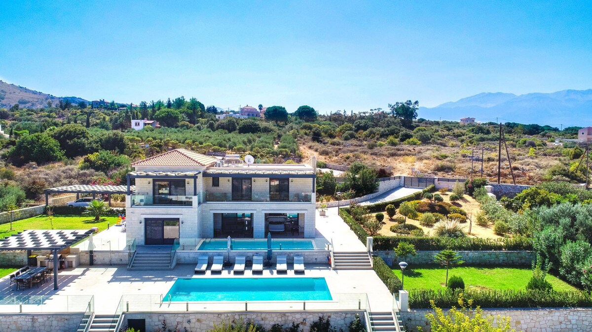 An aerial view of Villa Marevista is presented, showcasing a modern two-story structure with large windows and a spacious pool area. Surrounding gardens and landscaping enhance the villa's serene setting, complete with a sunlit pool, sunbeds, and lush greenery extending into the distance.