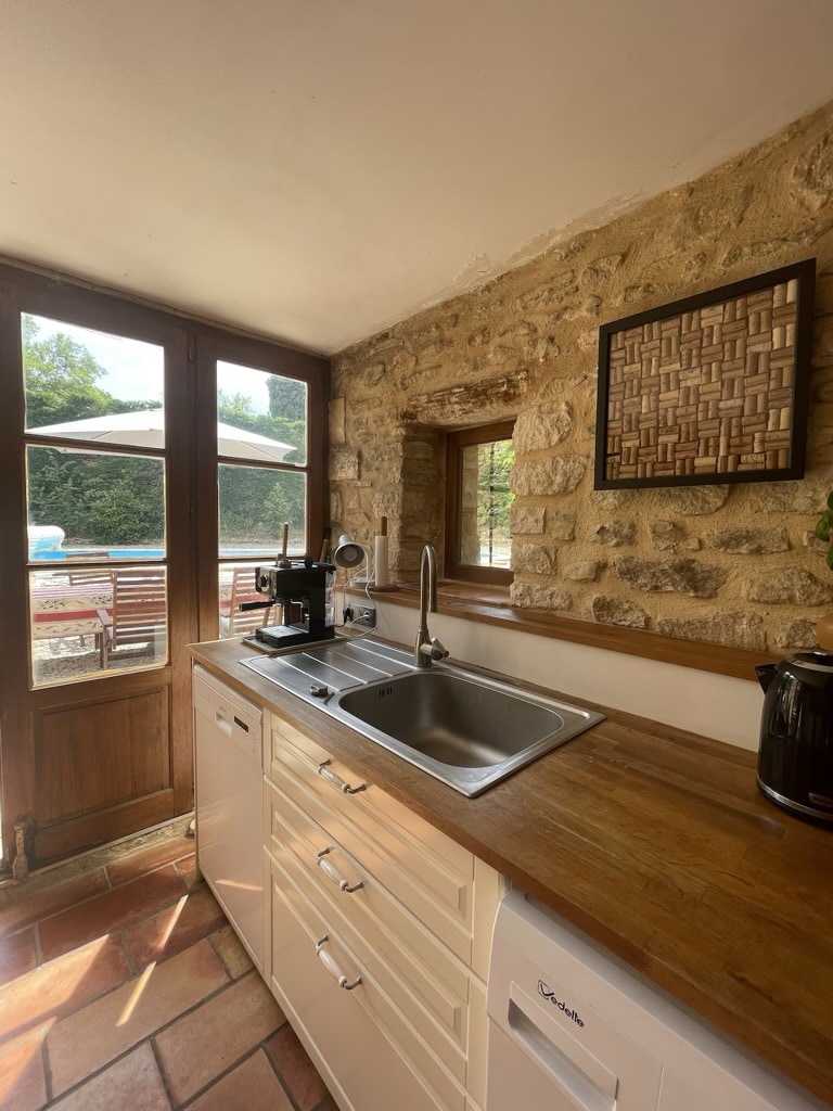 A well-equipped kitchen features a clean work surface with a stainless steel sink and a coffee machine. Natural light flows through the window, highlighting the stone wall and wooden cabinetry. A view of the outdoor terrace adds a sense of openness to the space.