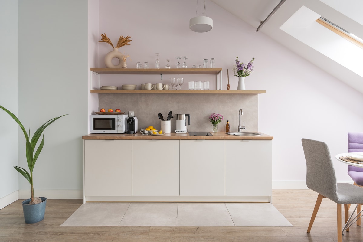 A fully equipped kitchenette features a light wood countertop and modern white cabinetry. Glassware and decorative items are displayed on shelving above. A microwave and coffee maker are included on the counter. A plant in a pot adds a touch of greenery to the space.