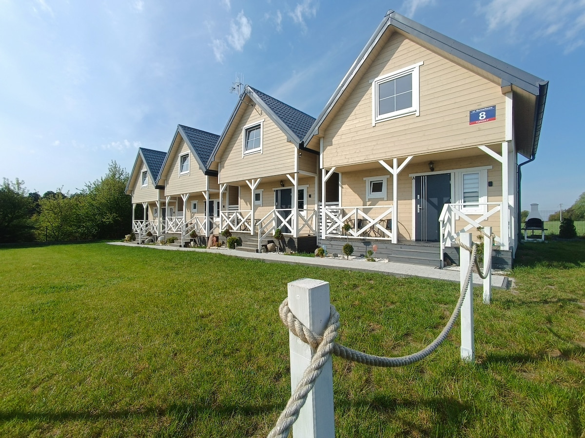 A row of newly constructed holiday homes is set against a clear blue sky. Each home features a sloped roof and a front porch with a railing. Lush green grass surrounds the buildings, enhancing the sense of spaciousness in the outdoor area.