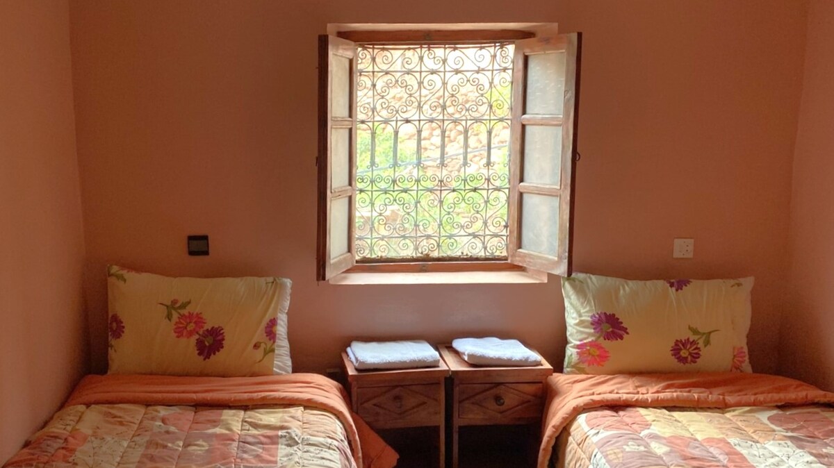 A cozy room with two comfortable beds adorned with floral-patterned linens. A window with intricate wrought iron grilles opens to views of the surrounding area. Nightstands are placed between the beds, each holding neatly stacked towels.
