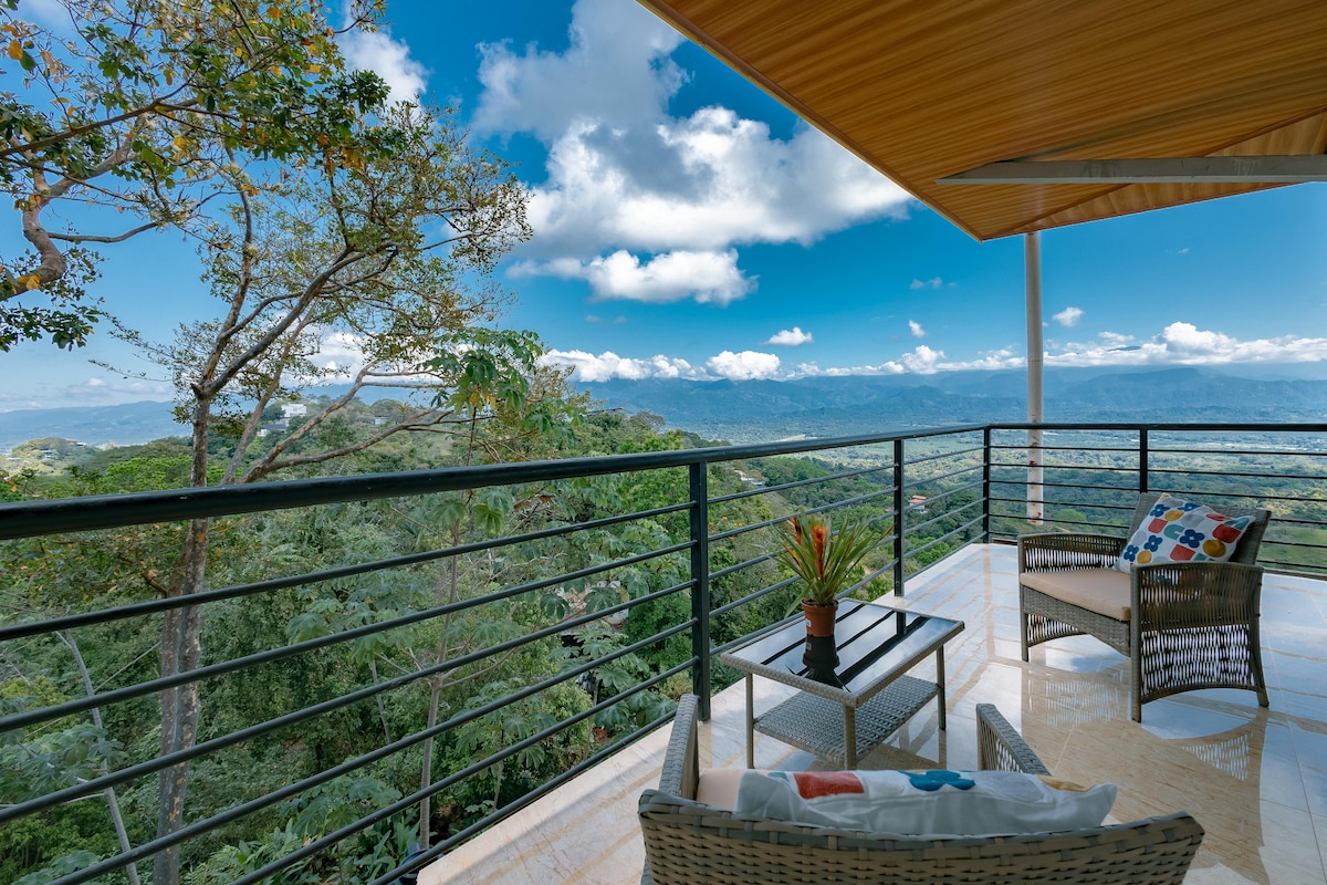 A balcony is presented with two chairs and a small table, overlooking a lush green landscape. The expansive view reveals mountains and a partly cloudy sky. A plant adds a touch of nature, while a decorative pillow brings color to the seating area.