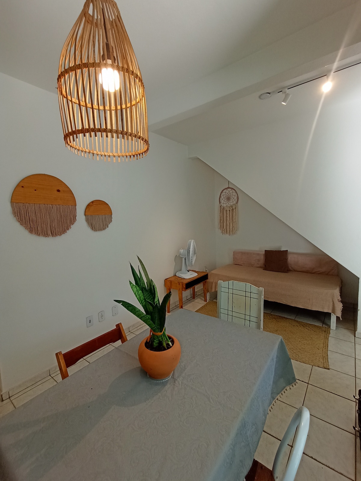 A dining area features a table set with a beige tablecloth, alongside four wooden chairs. A potted plant sits at the center of the table, while woven wall decorations add texture to the wall. A simple seating arrangement is visible in the background.