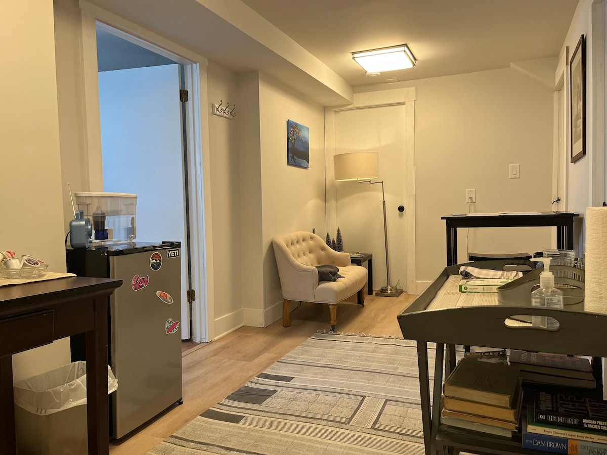 A well-lit hallway leads to a cozy seating area featuring a light-colored chair beside a lamp. A small table with books is present, and a compact refrigerator decorated with stickers is visible. The space is accentuated by a textured area rug and soft wall colors.