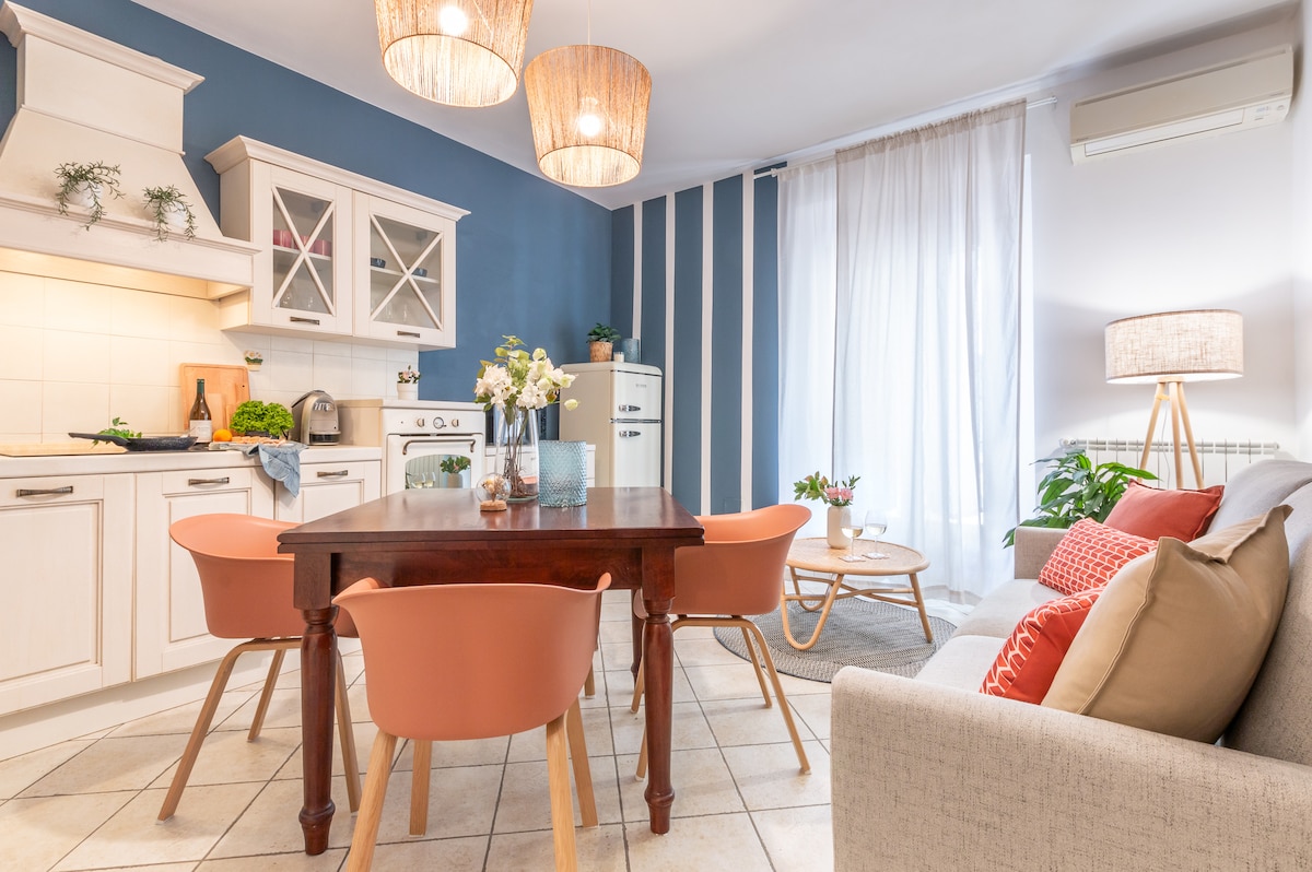 A modern kitchen and dining area is presented with light-colored cabinetry and a wooden dining table surrounded by four chairs. A cozy seating area features a gray sofa with colorful cushions. Natural light filters through sheer curtains, enhancing a welcoming ambiance.