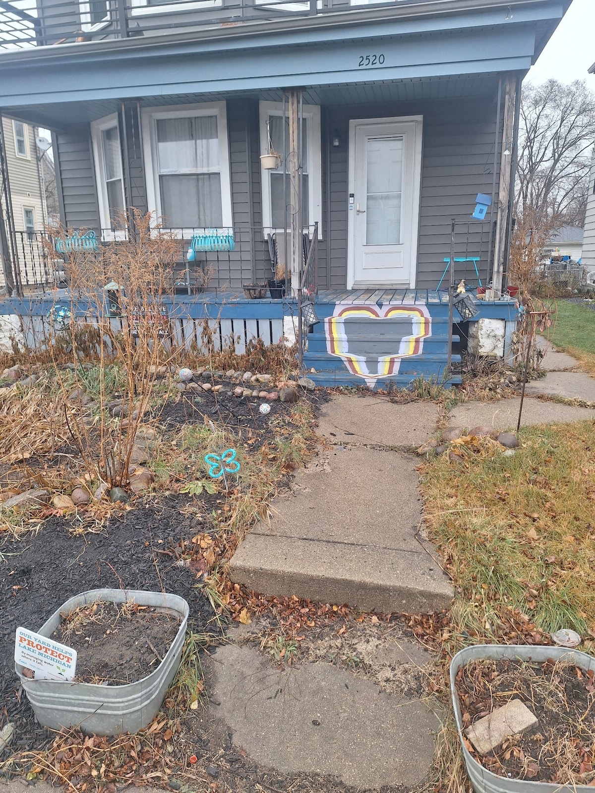 An inviting entryway features a heart design painted on the porch steps, surrounded by bare shrubs and decorative stones. A pathway guides visitors to the front door, with planters visible on either side, adding a personal touch to the home's facade.