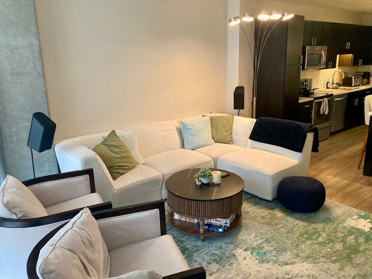 A stylish living area features a modern sectional sofa adorned with soft throw pillows. A round coffee table sits at the center, accompanied by two armchairs. A floor lamp provides ambient lighting, while the fully equipped kitchen is visible in the background.