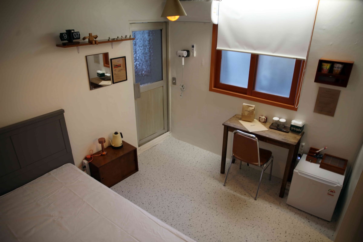 The room features a spacious bed and a wooden desk accompanied by a chair. A small refrigerator and amenities are visible against the wall. Natural light enters through a window fitted with a light-filtering shade, creating a bright and airy environment.