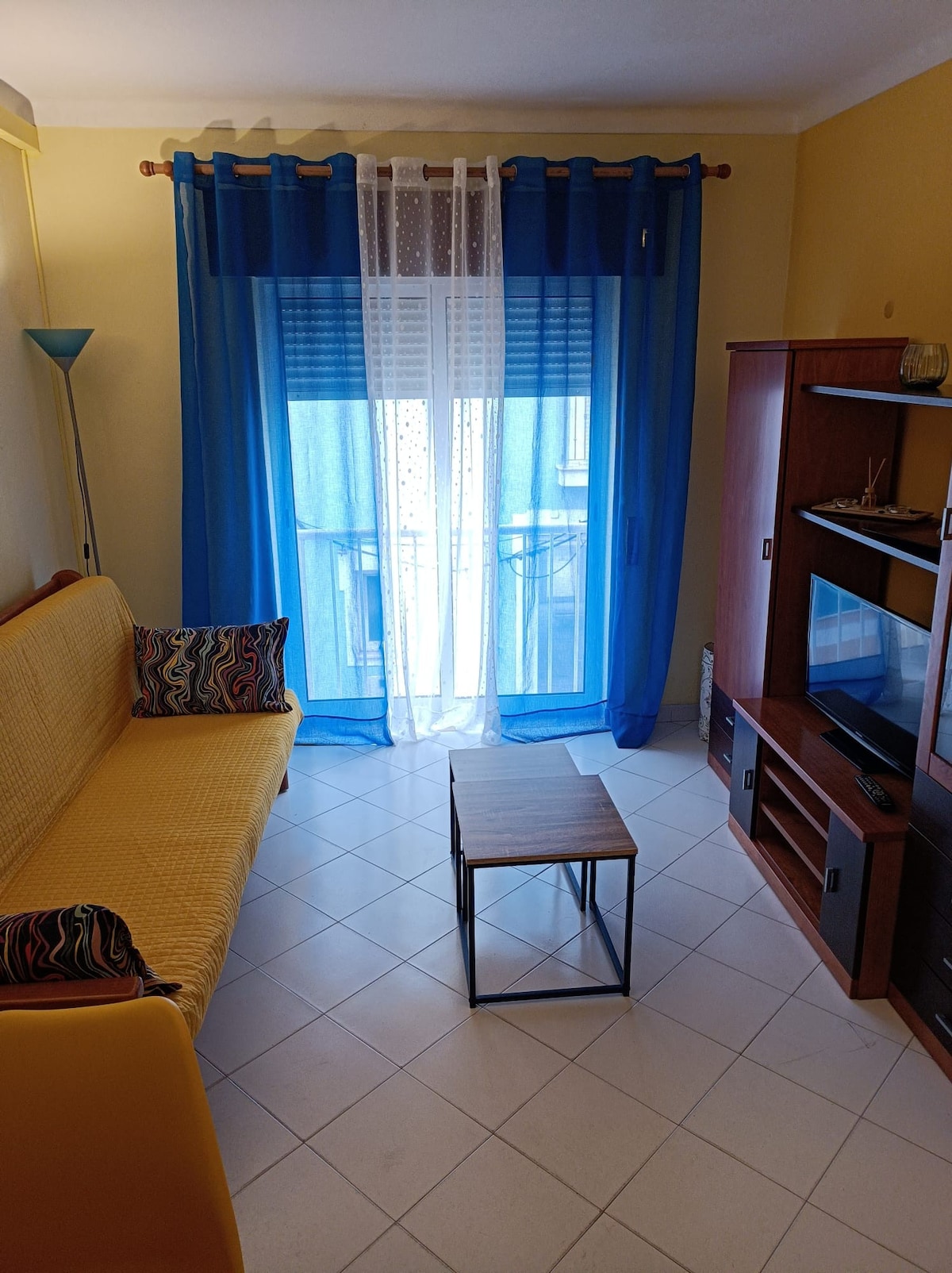 The living area features a yellow sofa bed adorned with patterned cushions. A small, minimalist coffee table sits in front, complemented by a wooden cabinet with a television. Sheer blue curtains allow natural light to fill the space, enhancing the airy feel of the room.