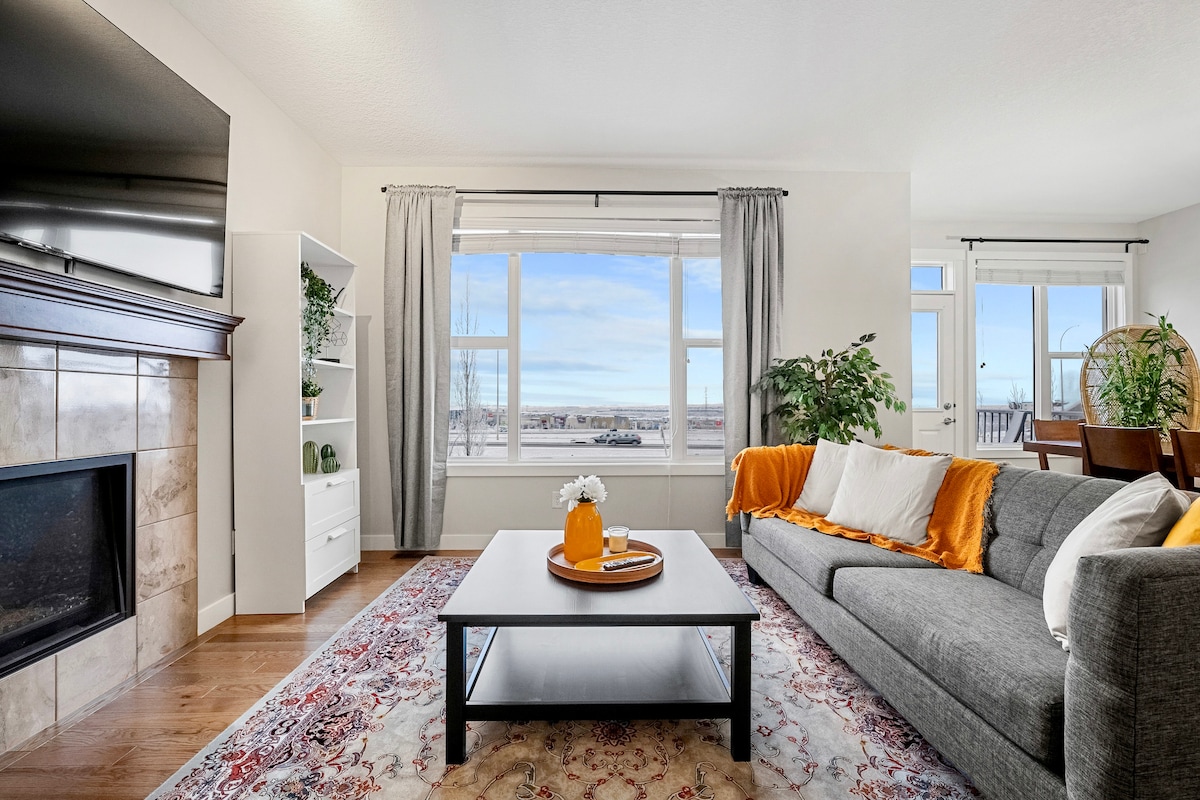 A welcoming living area features a gray sectional sofa adorned with orange and white throw pillows. A decorative coffee table centers the space, with a vase and fruit arrangement on top. Natural light floods in through large windows, highlighting the views and soft furnishings.