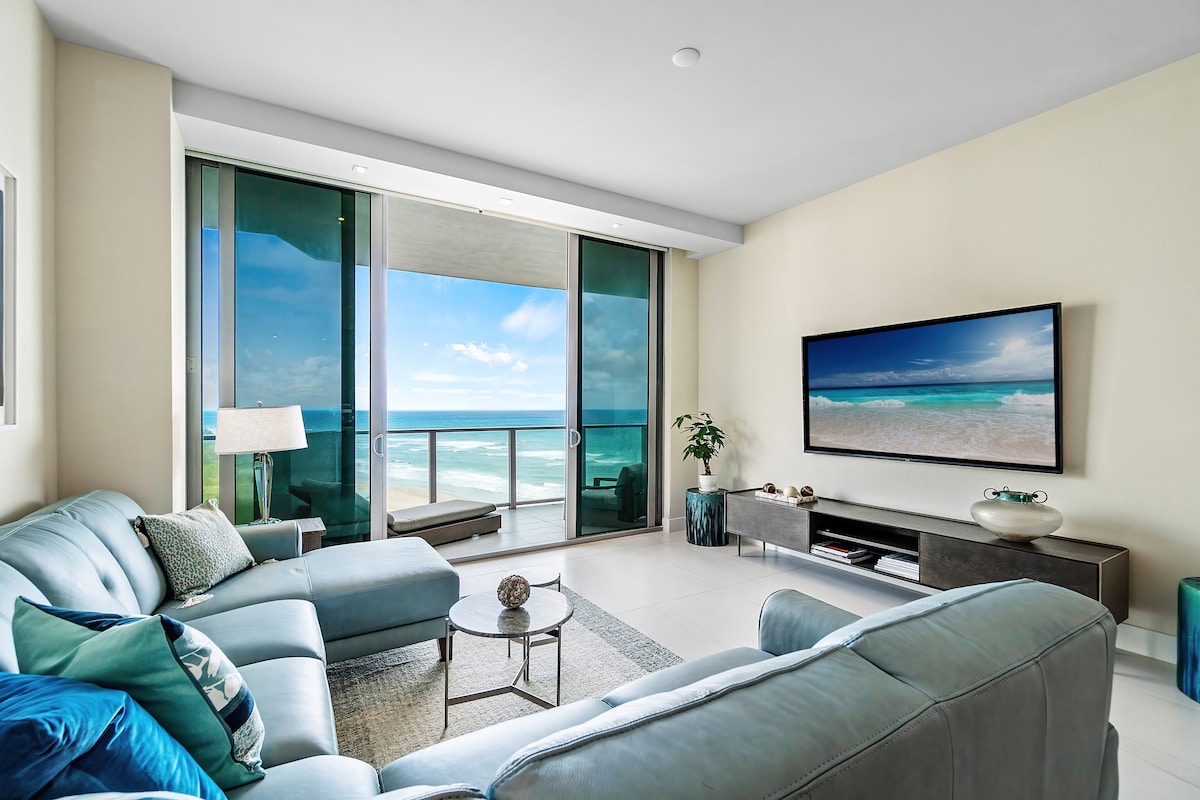 The living area features a light blue sectional sofa positioned near a glass balcony door, which reveals stunning ocean views. A large wall-mounted flatscreen TV complements the modern decor, with a coffee table placed centrally on the polished floor, contributing to the space's airy ambiance.