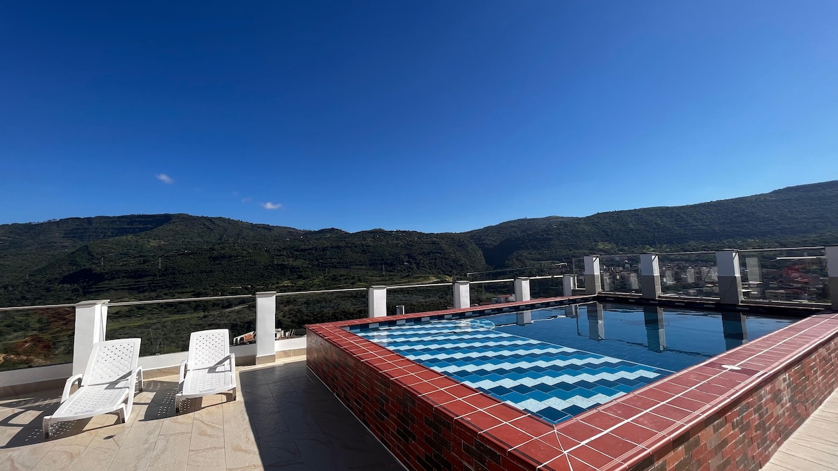 A rooftop pool area features a rectangular swimming pool with a distinct blue pattern, surrounded by a brick border. Two white lounge chairs are positioned nearby, and a clear blue sky stretches above, while lush green mountains are visible in the background.