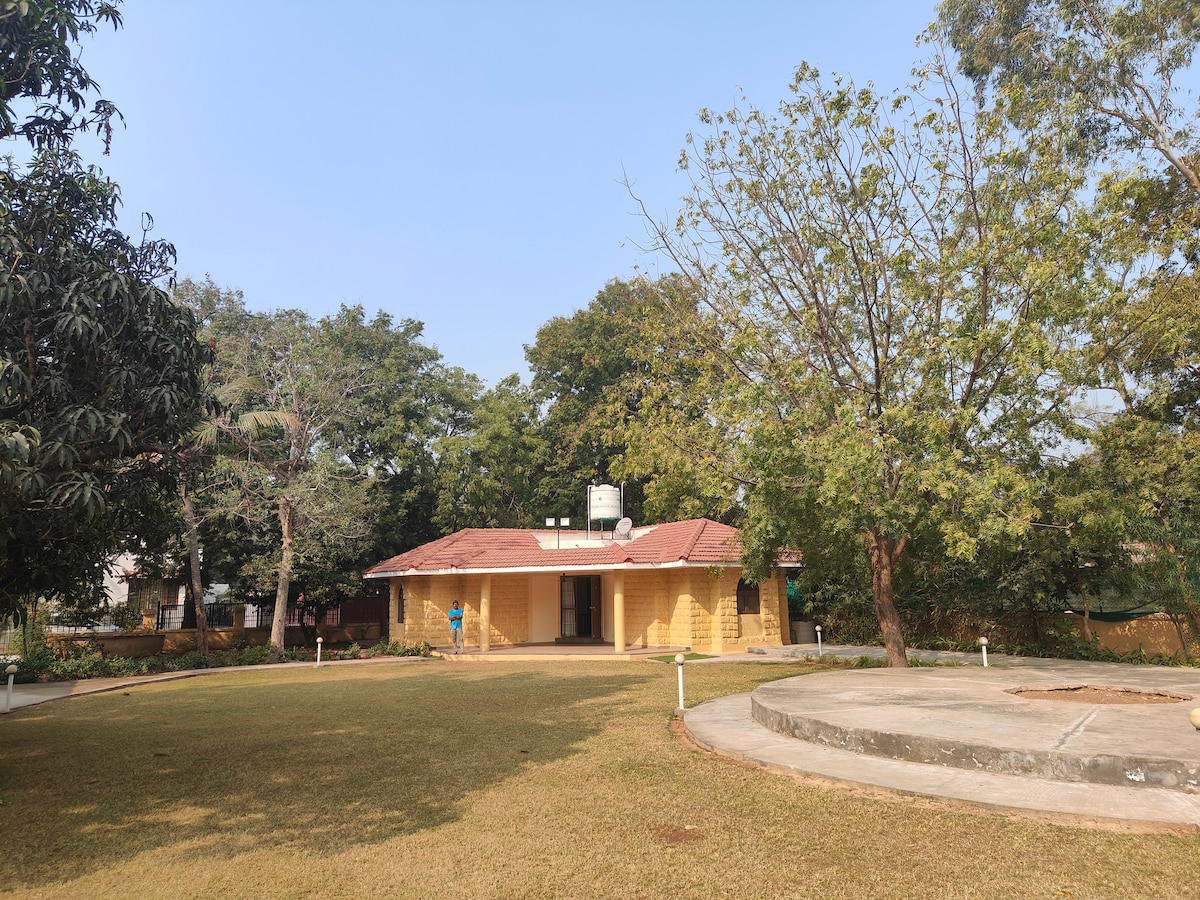 A single-story retreat is surrounded by lush greenery, featuring a charming, yellow-paneled exterior with a tiled roof. A spacious lawn leads to the entrance, and a circular stone area can be seen to the right, providing an open space for gatherings.