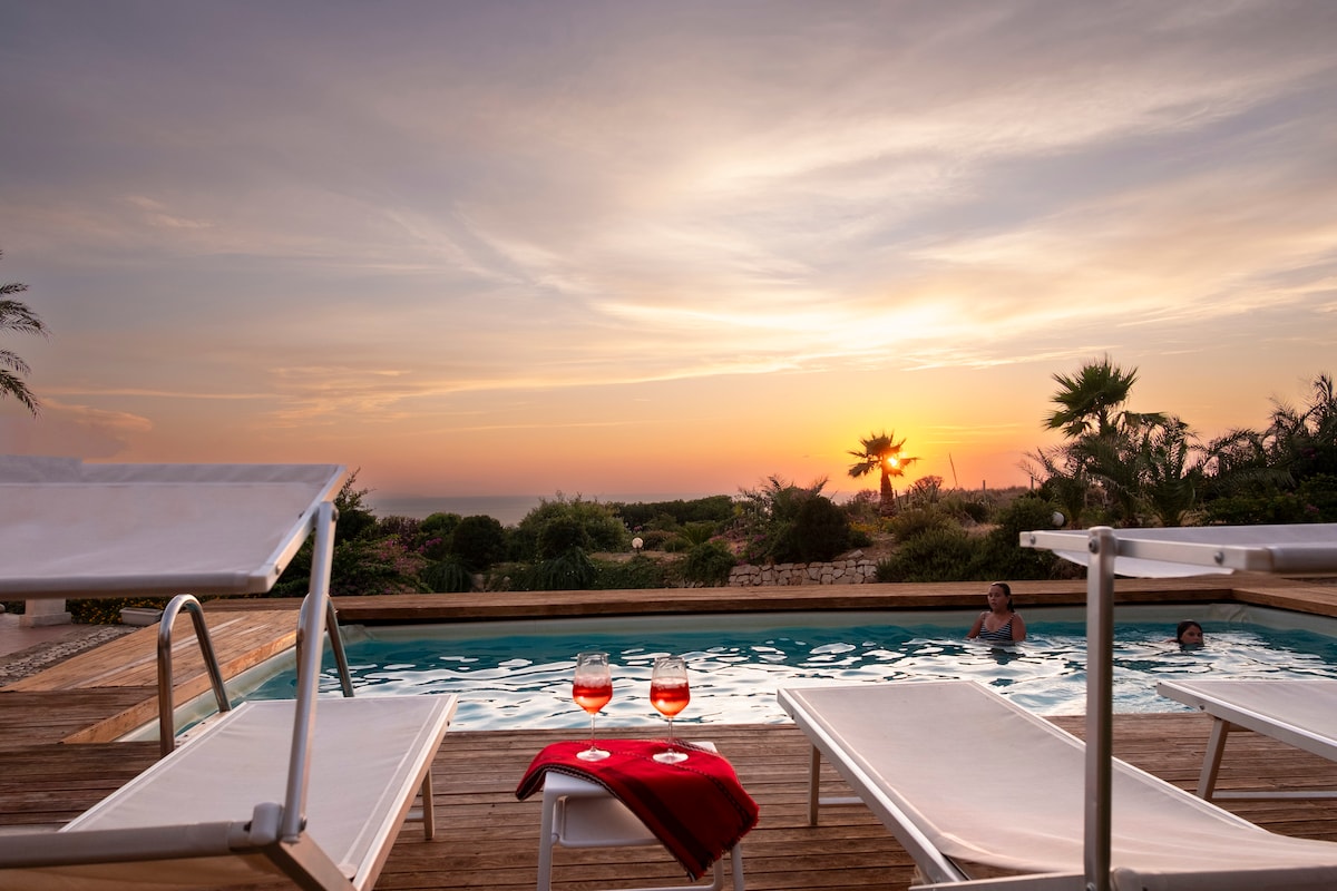 The image features a private pool surrounded by sun loungers, set against a vibrant sunset. Two glasses of drink are placed on a small table, creating a relaxing atmosphere. Lush greenery and palm trees can be seen in the background, enhancing the serene setting.
