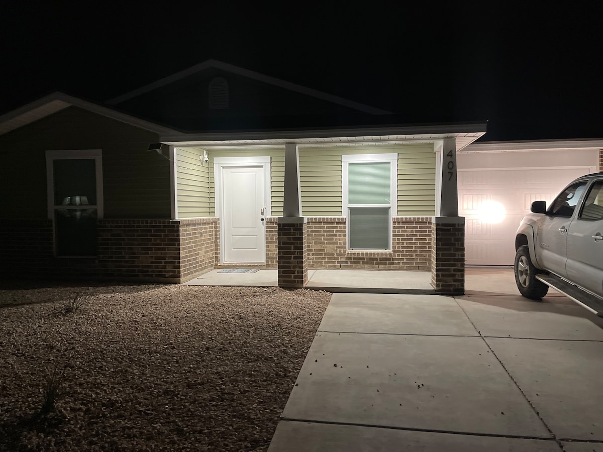 The exterior of a newly constructed home is illuminated at night, showcasing a well-lit entryway. A front porch is visible, featuring a single door and two windows. The driveway is spacious, with a parked vehicle nearby, set against a backdrop of soft landscaping.