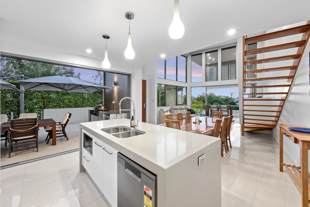 An open-plan kitchen and dining area are shown, featuring a sleek kitchen island with a sink and modern appliances. Large glass doors provide access to a covered outdoor dining space. Natural light illuminates the area, highlighting the contemporary design and spaciousness.